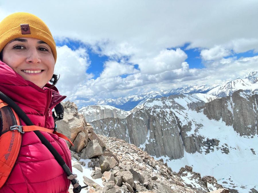 Sahar smiling high on Mount Whitney with snow-covered Sierra Nevada peaks behind her